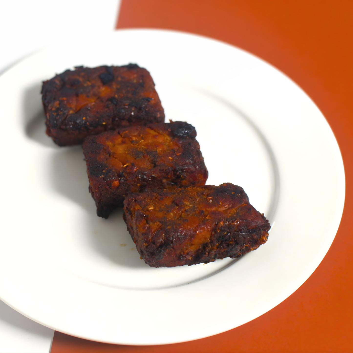 Three pieces of fried tempeh on a white plate with an orange background