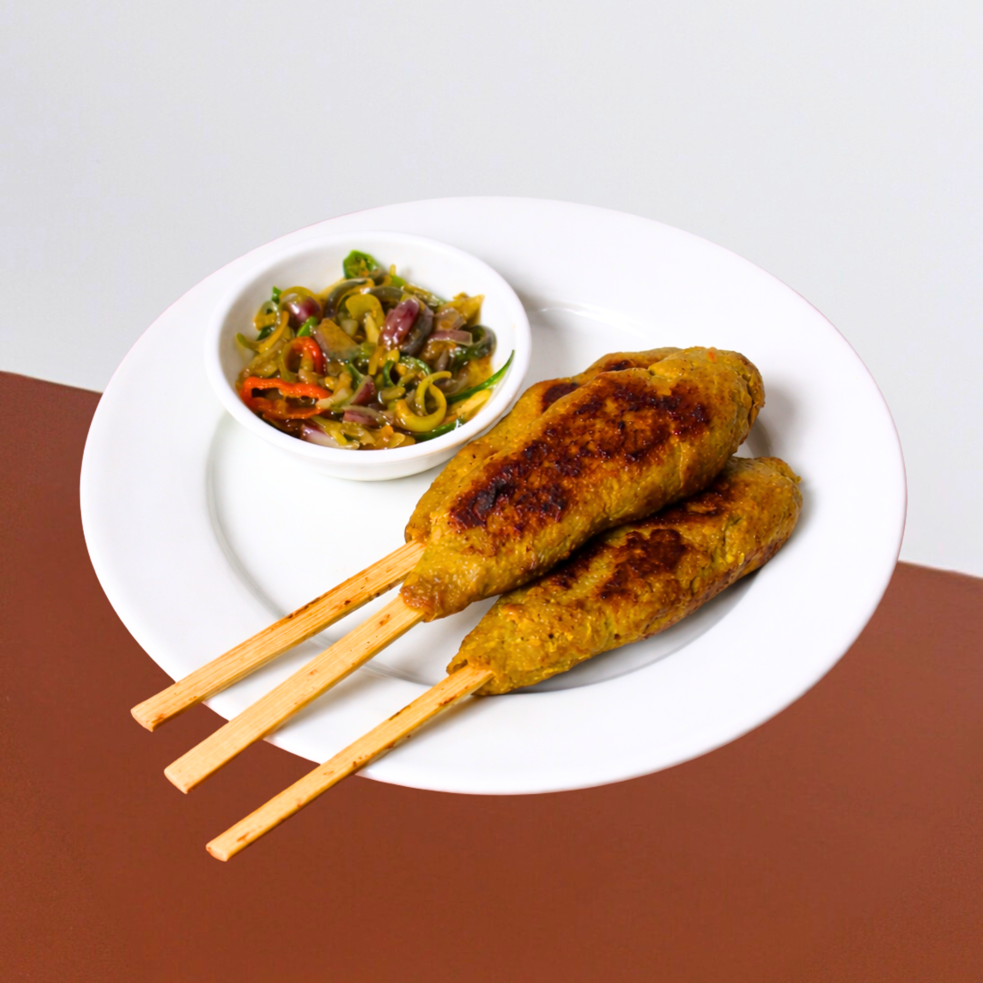 Three chicken skewers on a white plate with a side of colorful vegetables on a brown surface.